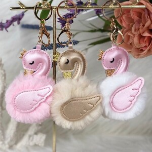 May include: Three fluffy swan keychains with gold accents. The swans are pink, white, and beige. The keychains are hanging on a gold stand.