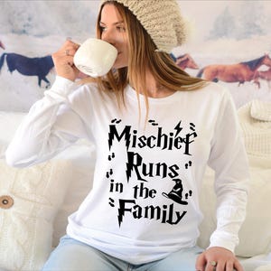 Peut inclure: T-shirt blanc à manches longues avec l'inscription noire "Mischief Runs in the Family". Le motif comprend un éclair, des empreintes de pas et un chapeau de sorcier. Le t-shirt est porté par une personne.