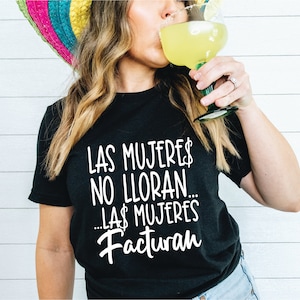 May include: Black t-shirt with white text that reads "LAS MUJERES NO LLORAN... LAS MUJERES Facturan." A person is drinking a margarita, wearing a colorful sombrero. The margarita is in a green-stemmed glass.