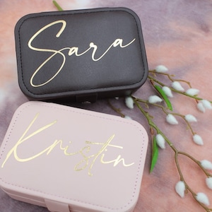 Könnte beinhalten: Zwei personalisierte Schmuckkästchen. Eines ist dunkelgrau mit dem Namen "Sara" in goldener Schrift, das andere ist hellrosa mit dem Namen "Kristin" in goldener Schrift. Beide sind rechteckig mit einem Schnappverschluss.
