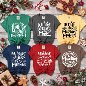 May include: Six short-sleeved t-shirts in green, dark grey, yellow, navy blue, red, and brown, each featuring white graphic text and holiday-themed illustrations including reindeer, gingerbread men, Santa's sleigh, and lightning bolts.
