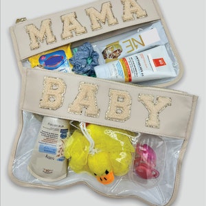 Può includere: Una pochette beige con cerniera e pannelli in vinile trasparente. La pochette ha le parole "MAMA" e "BABY" in lettere dorate con glitter. La pochette contiene vari articoli per la cura del bambino, tra cui un giocattolo da bagno a forma di anatra gialla, un ciuccio rosa e una bottiglia di shampoo per bambini.