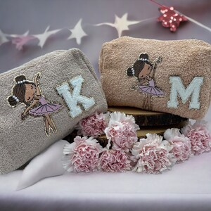 May include: Two beige and brown fabric cosmetic bags with embroidered ballerina figures and the letters K and M. The bags are on a wooden surface with pink carnations.
