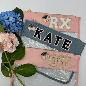 May include: Three clear zippered pouches with pink and blue fabric exteriors. Each pouch has a different name on it: "RX", "KATE", and "OY". Each pouch also has a gold graduation cap pin with the word "GRADUATE" on it.