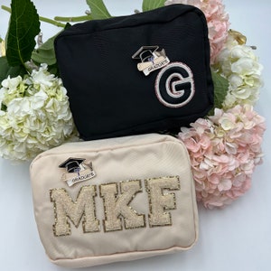May include: Two cosmetic bags, one black and one beige. The black bag has a gold graduation cap and diploma charm with the text "Graduate" and a gold letter "G". The beige bag has a gold graduation cap and diploma charm with the text "Graduate" and the text "MKF" in gold glitter letters.