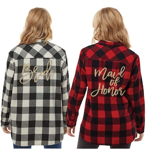 May include: Two flannel shirts with a black and white plaid pattern and a red and black plaid pattern. The back of the black and white shirt says "Bride" in gold glitter. The back of the red and black shirt says "Maid of Honor" in gold glitter.