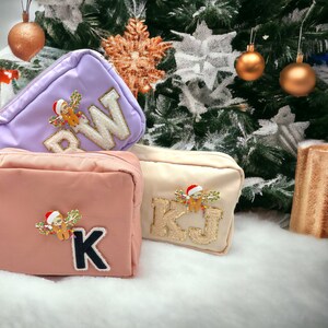 May include: Three personalized cosmetic bags with a Christmas theme. The bags are purple, pink, and beige. Each bag has a sloth wearing a Santa hat and holding Christmas lights. The bags are monogrammed with the letters "RW", "K", and "KJ".