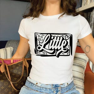 May include: White cropped t-shirt featuring a black graphic of the word "Little" in a stylised font with hearts around it.