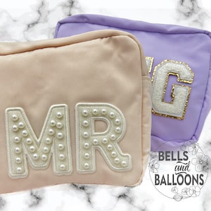 May include: Two pink and purple zippered pouches with pearl-embellished letters spelling "MR" and "G". The pouches are on a marble surface. The text "BELLS and BALLOONS" is visible in the lower right corner of the image.