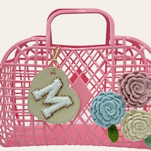 May include: Pink plastic tote bag with a heart-shaped charm with a sparkly letter "N" and three crocheted flowers in pink, blue, and cream.