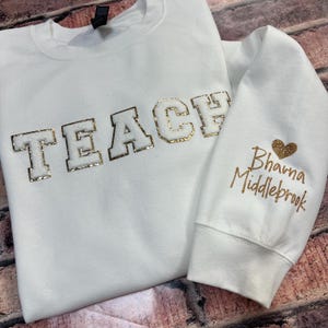 May include: A white sweatshirt with the word "TEACH" in gold glitter letters across the chest. The right sleeve has the name "Bhavna Middlebrook" in gold glitter with a gold glitter heart.