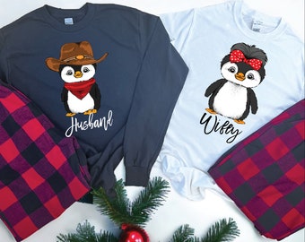 May include: A dark gray long-sleeve shirt with a penguin wearing a cowboy hat and red bandana, labeled Husband, and a white t-shirt with a penguin wearing a red polka-dot bow, labeled Wifey, both paired with red and black plaid pajama pants.