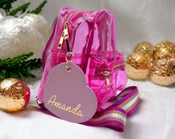 May include: A pink clear plastic backpack with a heart-shaped tag that says "Amanda". The backpack has a gold zipper and a pink and white striped strap.