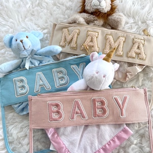 May include: Three clear zippered pouches with plush toys and blankets inside. The pouches have the words "MAMA", "BABY", and "BABY" embroidered on them in gold, blue, and pink thread.