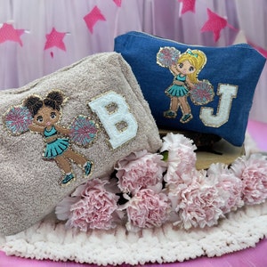 Peut inclure: Deux pochettes en tissu personnalisées avec des pom-pom girls et des lettres brodées. La pochette de gauche est beige avec la lettre "B" et une pom-pom girl en uniforme bleu et blanc. La pochette de droite est bleue avec la lettre "J" et une pom-pom girl en uniforme bleu et blanc.