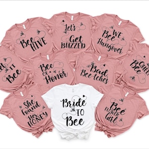 May include: Ten pink t-shirts with black text and bee graphics. The shirts have different sayings, including "Let it Bee", "Bee Hive", "Bee of Honor", "Bad Bee-tches", "Queen Bee", "We Bee Hungover", "Let's Get Buzzed", "She Found Her Honey", "Bride to Bee", and "Bee Flower Girl".