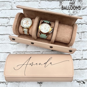 May include: A pink watch box with a personalized inscription that reads "Amanda". The box has three slots for watches and is open to reveal two watches inside. The box is on a white brick background.