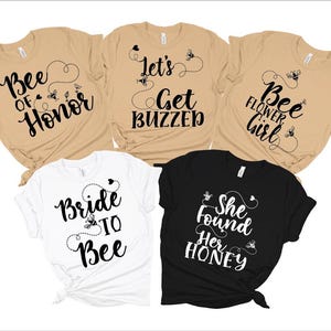 May include: Five beige t-shirts with black text and bee graphics. The shirts read "Bee of Honor", "Let's Get Buzzed", "Bee Flower Girl", "Bride to Bee", and "She Found Her Honey".
