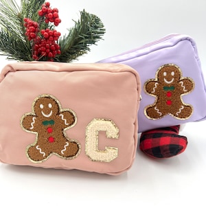 Peut inclure: Deux petits sacs cosmétiques rectangulaires. L'un est rose clair avec un patch de bonhomme en pain d'épices et un "C" doré à paillettes. L'autre est lavande avec un patch de bonhomme en pain d'épices. Décorations de Noël en arrière-plan.