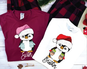 May include: Matching Christmas pajama sets featuring penguin graphics. One burgundy shirt with a pink Santa hat and the word "Sister", and a white shirt with a red Santa hat and the word "Brother". Plaid pajama pants complete the set.