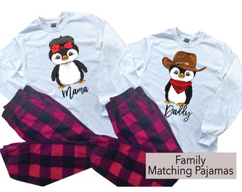 May include: Matching family pajamas set featuring white long-sleeve shirts with penguin graphics. One penguin wears a bow, the other a cowboy hat. Plaid pajama pants in red and black complete the set. Text reads "Mama" and "Daddy".