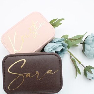 Könnte beinhalten: Zwei kleine, rechteckige Schmuckkästchen mit goldenen Schriftzügen. Die obere Schachtel ist rosa mit dem Namen "Kristen" in goldener Schrift. Die untere Schachtel ist braun mit dem Namen "Sara" in goldener Schrift.