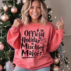 Peut inclure: Un sweat-shirt à col rond de couleur corail avec le texte noir Official Holiday Mischief Maker. Le motif comprend un éclair, des flocons de neige, un bonhomme en pain d'épices et un graphique de baguette magique intégré aux lettres.