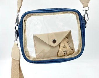 Puede incluir: Un bolso transparente de plástico con correa cruzada y ribete de denim azul. El bolso tiene una correa de tela beige y un bolsillo de tela beige con una letra "A" de purpurina dorada en la parte delantera.