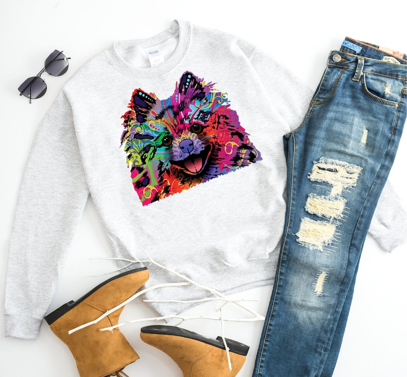 May include: A light gray sweatshirt featuring a vibrant, colorful graphic of a dog's face. The dog's fur is depicted in a kaleidoscope of colors. The image also includes blue jeans, brown boots, and sunglasses.