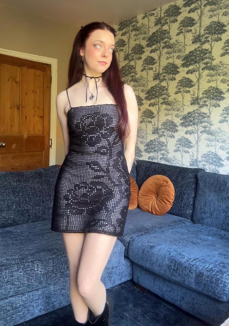 Op de afbeelding: Een zwarte mini-jurk met een bloemenpatroon. De jurk heeft dunne bandjes en een vierkante halslijn. Het is gemaakt van een gehaakt materiaal. De persoon draagt een zwarte choker en zwarte laarzen.