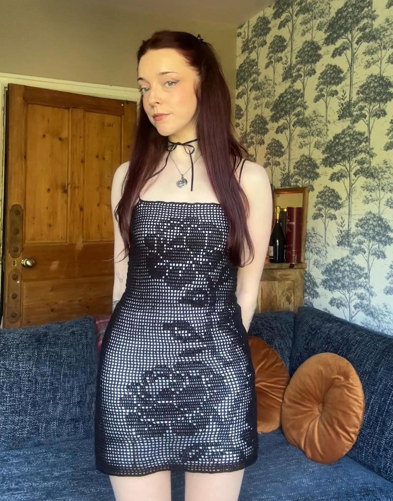 Op de afbeelding: Een zwart-witte gehaakte jurk met een bloemenpatroon. De jurk heeft een vierkante halslijn en spaghettibandjes. Het model draagt een zwarte choker-ketting met een zilveren hanger. De achtergrond omvat een blauwe bank en behang met patronen.