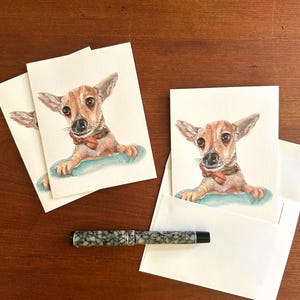 May include: Greeting cards featuring a watercolor painting of a small brown dog with a red bow tie and a green and red collar. The cards are on a wooden surface, with a pen and envelopes.