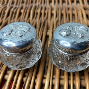 May include: Two small glass jars with silver lids. The lids are embossed with a design of three cherubs' heads.