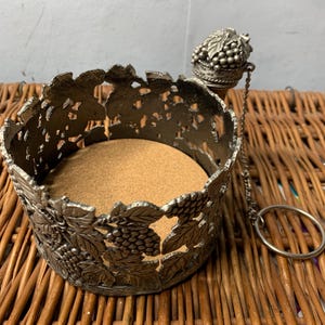 May include: A silver-colored wine bottle coaster with a grape and leaf design. The coaster has a cork base and a decorative grape cluster bottle stopper attached by a chain. The stopper has a ring for easy handling.