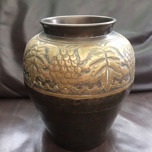Könnte beinhalten: Eine dunkelbraune und goldene Metallvase mit einem geprägten Design aus Trauben und Blättern.