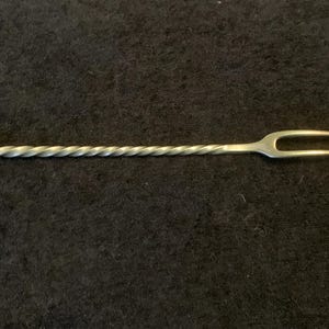 Könnte beinhalten: Ein silberner Barlöffel mit einem gedrehten Stiel und einer kleinen Kugel an einem Ende. Das andere Ende hat eine zweizinkige Gabel. Der Barlöffel ist zum Mischen von Cocktails und anderen Getränken bestimmt.