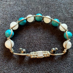 May include: A bracelet made with white and turquoise beads, with a silver clasp and black beads on the ends of the cord.