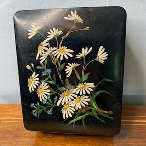 May include: A black rectangular box with a floral design. The box is decorated with white daisies and green leaves on a black background.