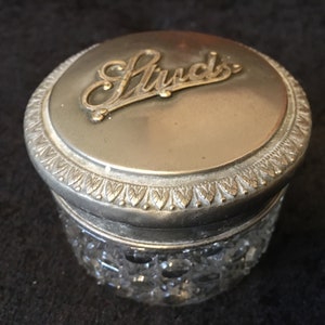 May include: A silver and glass jar with a lid that has the word "Studs" embossed on it.