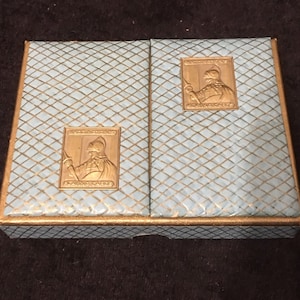 Könnte beinhalten: Eine blaue und goldene Spielkartenbox mit einem goldenen, geprägten Bild eines Ritters auf jeder Seite. Die Box hat ein Diamantenmuster.