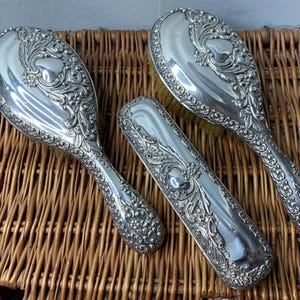 Könnte beinhalten: Ein Set aus drei versilberten Haarpflegewerkzeugen, bestehend aus einer Haarbürste, einem Kamm und einem Bürstenetui. Die Werkzeuge sind mit floralen Mustern verziert.