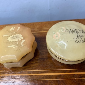 May include: Two round, white alabaster trinket boxes with floral designs. One box has a floral design and the other has the text "With Love from Eilat".