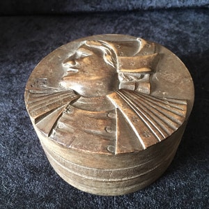 May include: A round wooden box with a carved portrait of a person wearing a hat. The box is made of dark wood and has a simple design.