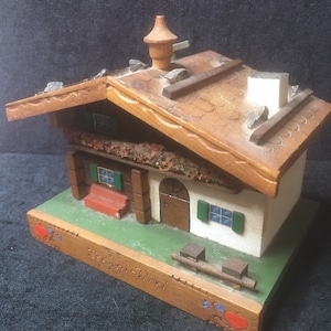 Könnte beinhalten: Ein hölzernes Miniatur-Chalet mit braunem Dach, weißen Wänden und grünen Fensterläden. Der Sockel ist braun mit dem Text "Rechal-Tirol" und dekorativen Herzen. Das Dach hat einen Schornstein und ist mit kleinen Steinen verziert.
