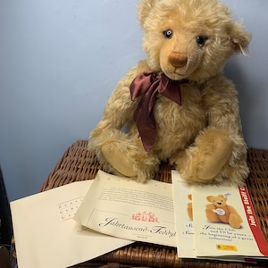 May include: A light brown teddy bear with a brown bow tie sitting on a wicker basket. The bear is a Steiff brand and is part of the Jahrtausend-Teddyt collection. There are two pieces of paper in front of the bear, one with the text "Jahrtausend-Teddyt" and the other with the text "Join the Steiff Club".