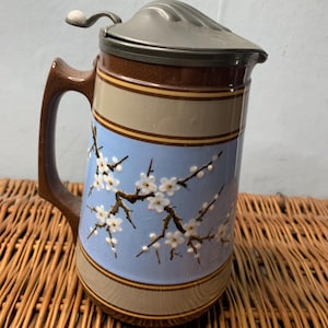 May include: A vintage ceramic beer stein with a hinged metal lid. The stein has a brown handle and base, with a light blue central band decorated with white floral branches. The top and bottom bands are tan and brown.