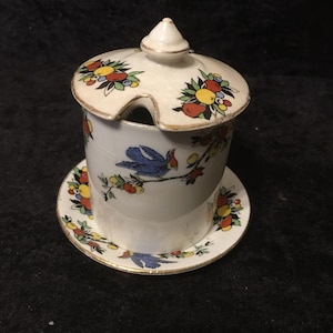 May include: A white ceramic sugar bowl with a lid and a matching saucer. The bowl and saucer are decorated with a floral pattern featuring blue birds, red and yellow fruit, and green leaves. The lid has a gold trim.