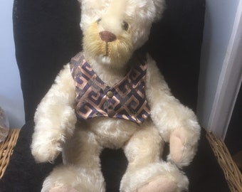 May include: A white teddy bear wearing a brown and purple patterned vest. The bear is sitting on a black chair.