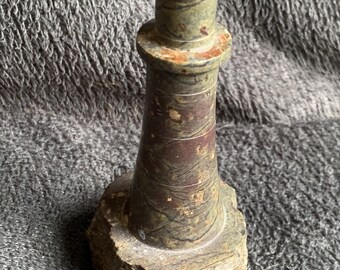 Puede incluir: Pequeña escultura de faro de piedra. El faro es principalmente verde y marrón con una superficie texturizada. La base es una piedra natural rugosa. La parte superior del faro tiene una pequeña tapa redondeada.