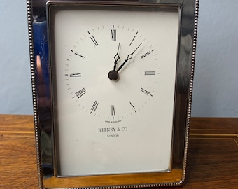 May include: Silver-plated mantel clock with Roman numeral hour markers and a white face. The clock has a beveled glass face and a silver-plated frame with a beaded edge. The clock face reads "Kitney & Co. London" and "Made in England".